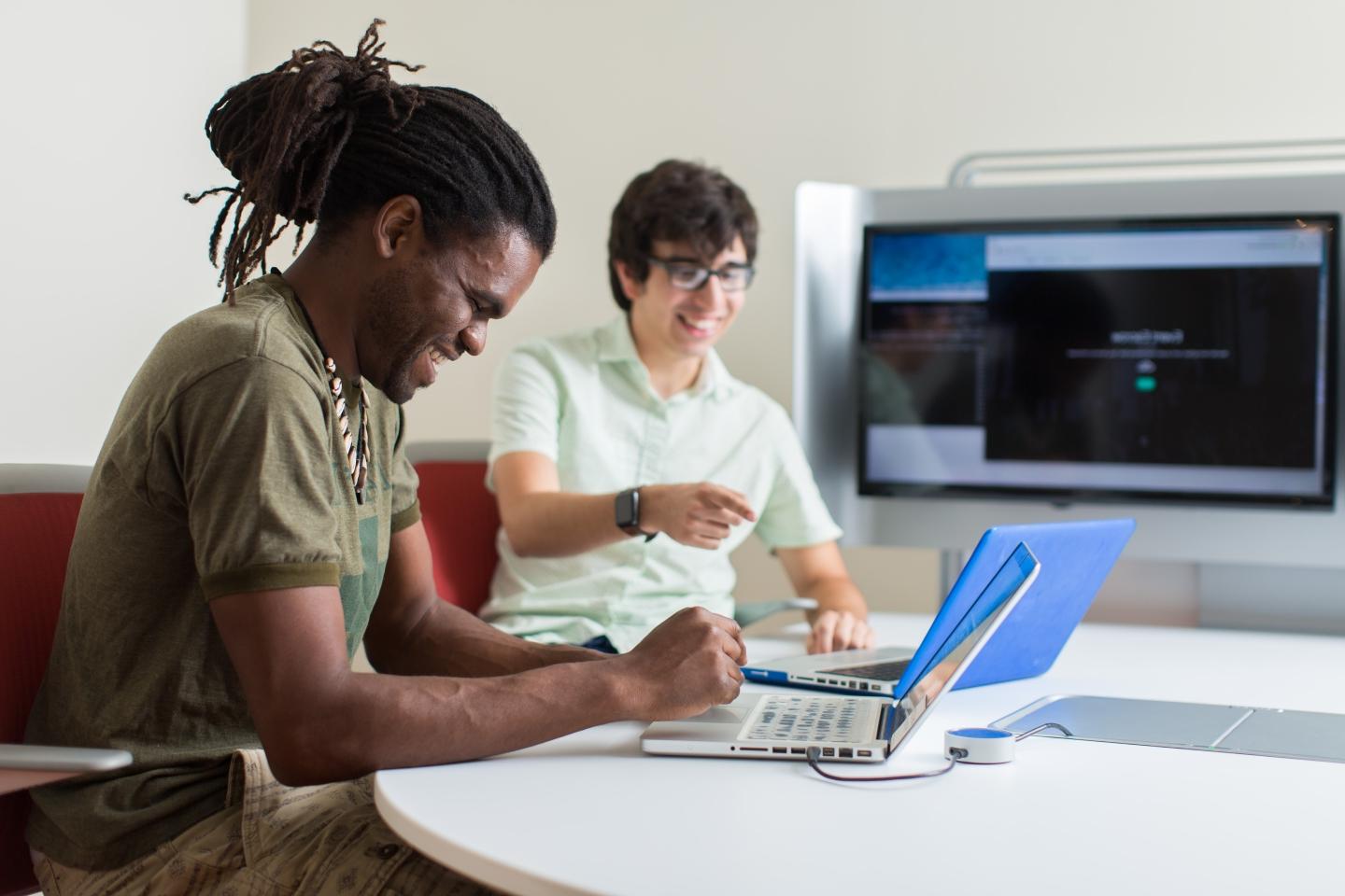 Two students talk and laugh while working on their laptops.